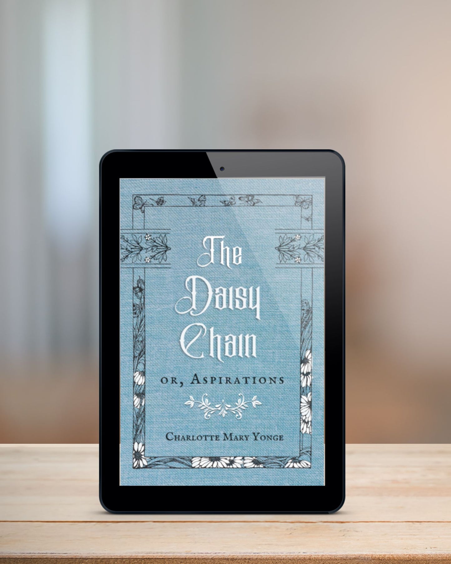 Black tablet displaying ebook of the pale blue and white cover of "The Daisy Chain or, Aspirations." It stands upright on a blonde table in front of a blurred background.