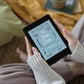 A small black tablet being held by a white woman's hands in a "cozy vibe" with sweater, blankets, mug of tea nearby. The pale blue and white cover of the ebook "The Daisy Chain or, Aspirations" adds to the charm.