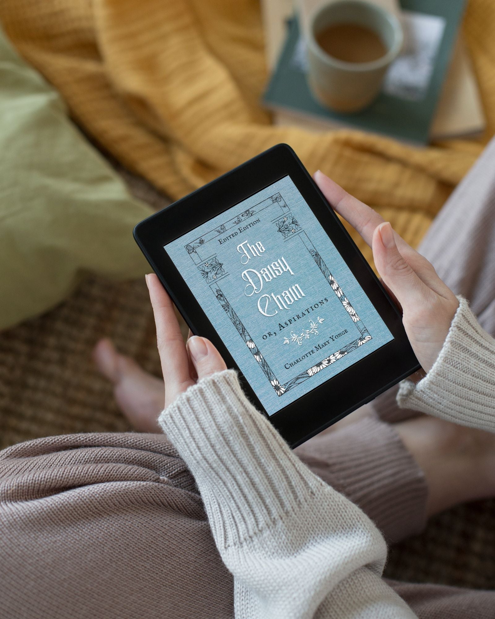 A small black tablet being held by a white woman's hands in a "cozy vibe" with sweater, blankets, mug of tea nearby. The pale blue and white cover of the ebook "The Daisy Chain or, Aspirations" adds to the charm.