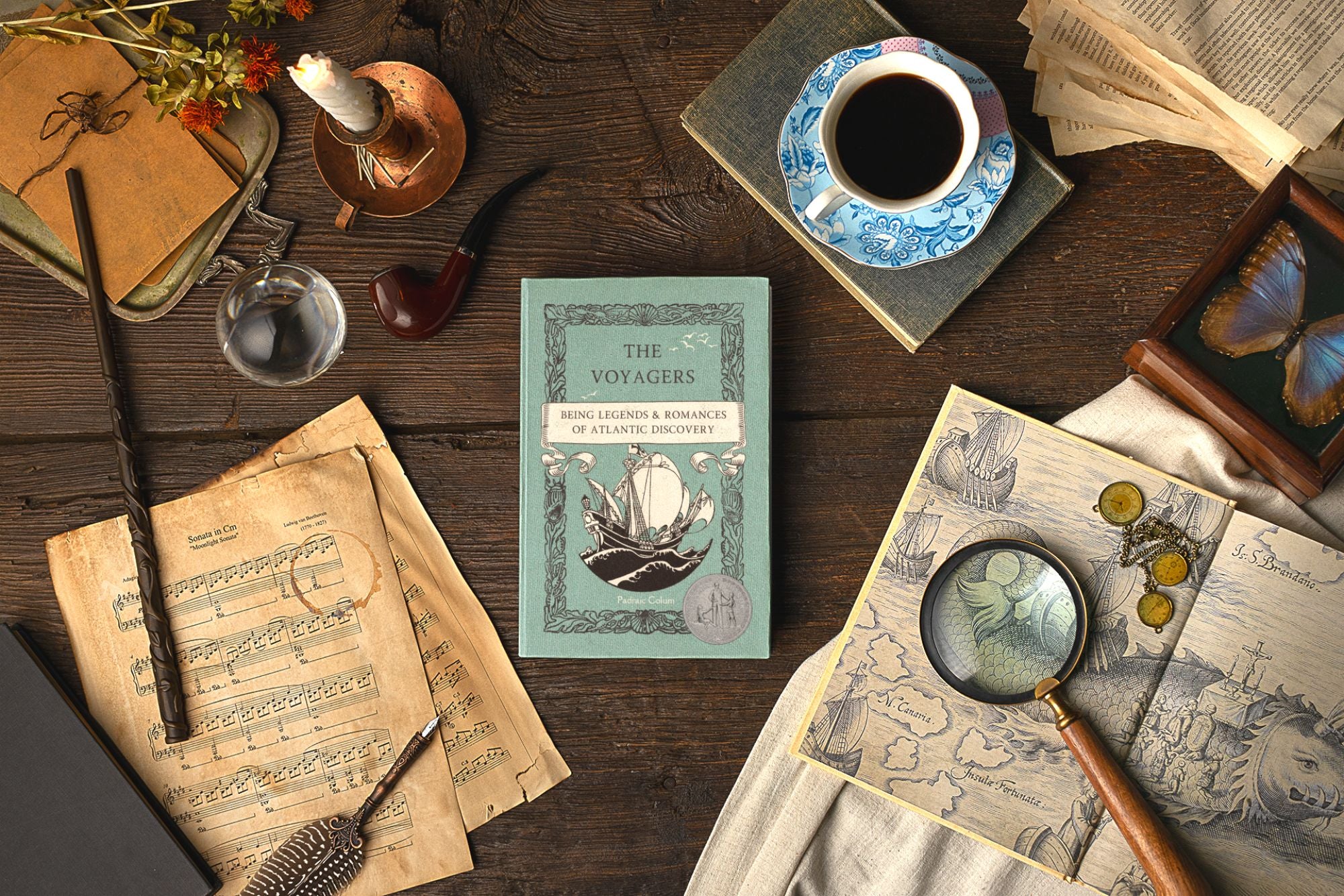 A dark wooden table holds old papers, vintage books, a blue china teacup and saucer, and a feather pen near a candle and a magic wand. In the center is The Voyagers by Padraic Colum.