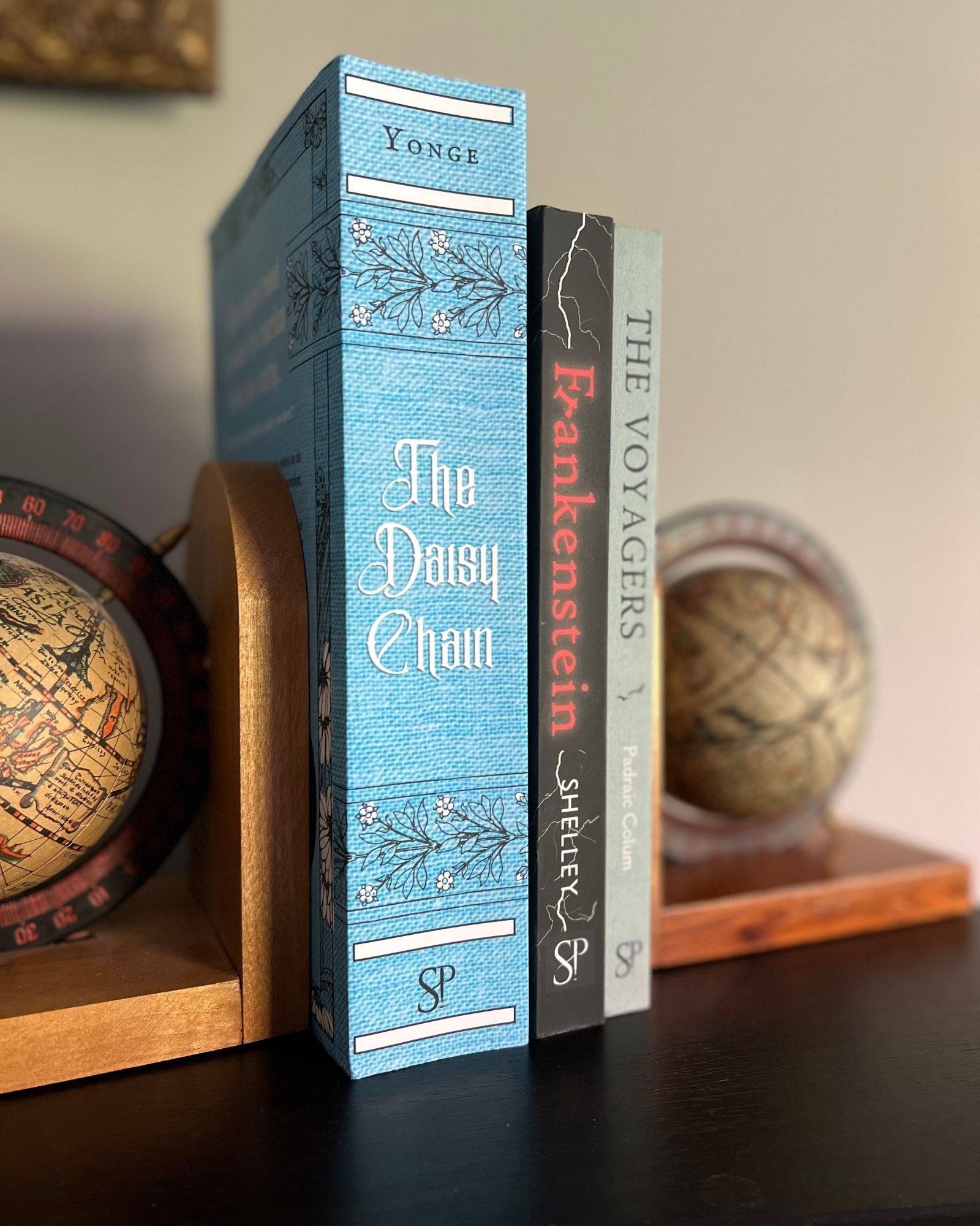 Globe bookends hold the paperback editions of Smidgen Press books, spines facing viewer. From left to right, "The Daisy Chain," "Frankenstein", and "The Voyagers".