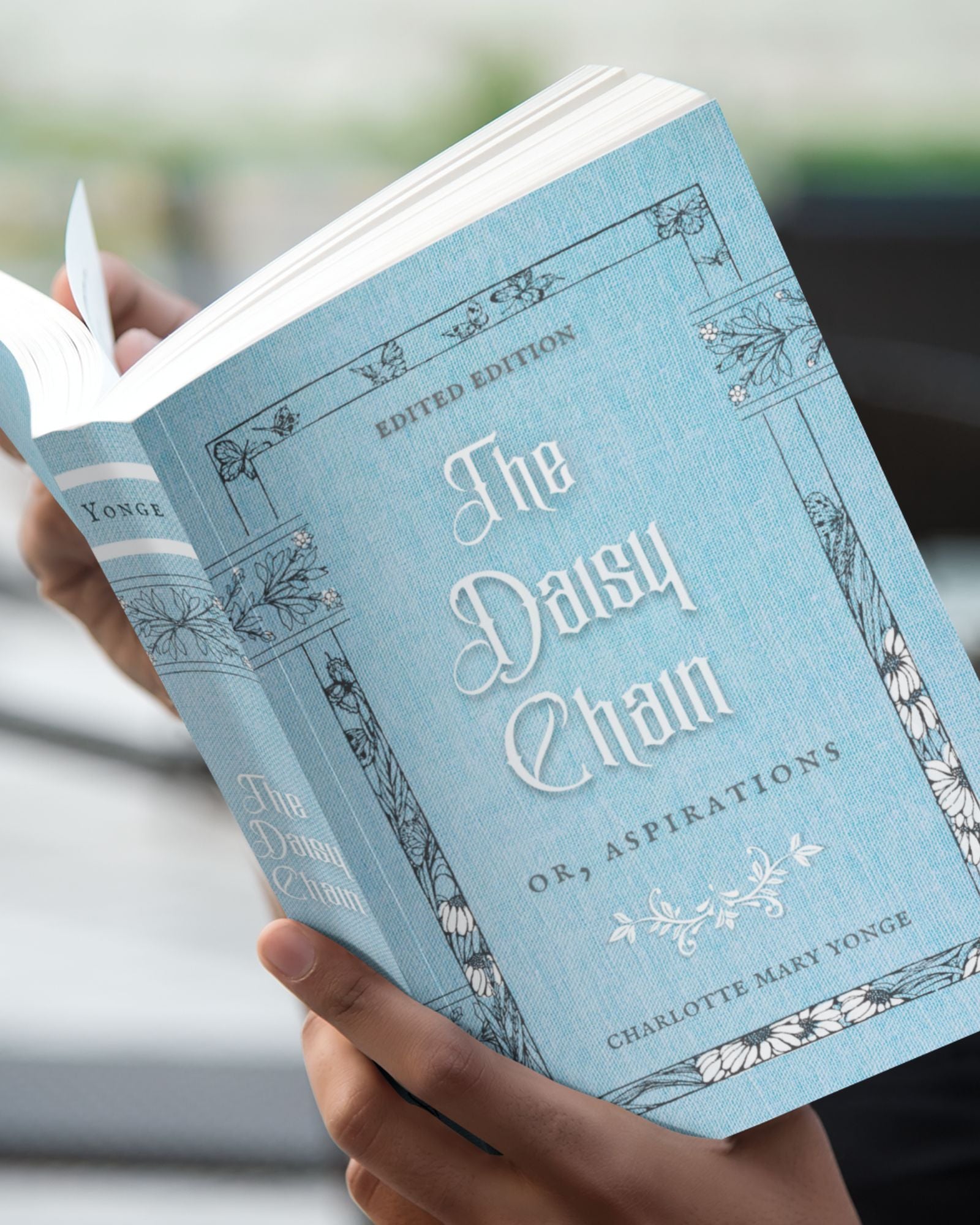 The paperback edition of The Daisy Chain or, Aspirations is being held open by white hands in front of a blurred background.