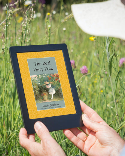 A small black tablet is being held by white hands in front of a field of wildflowers by a person wearing a white brimmed hat. On display is the golden cover of "The Real Fairy Folk".