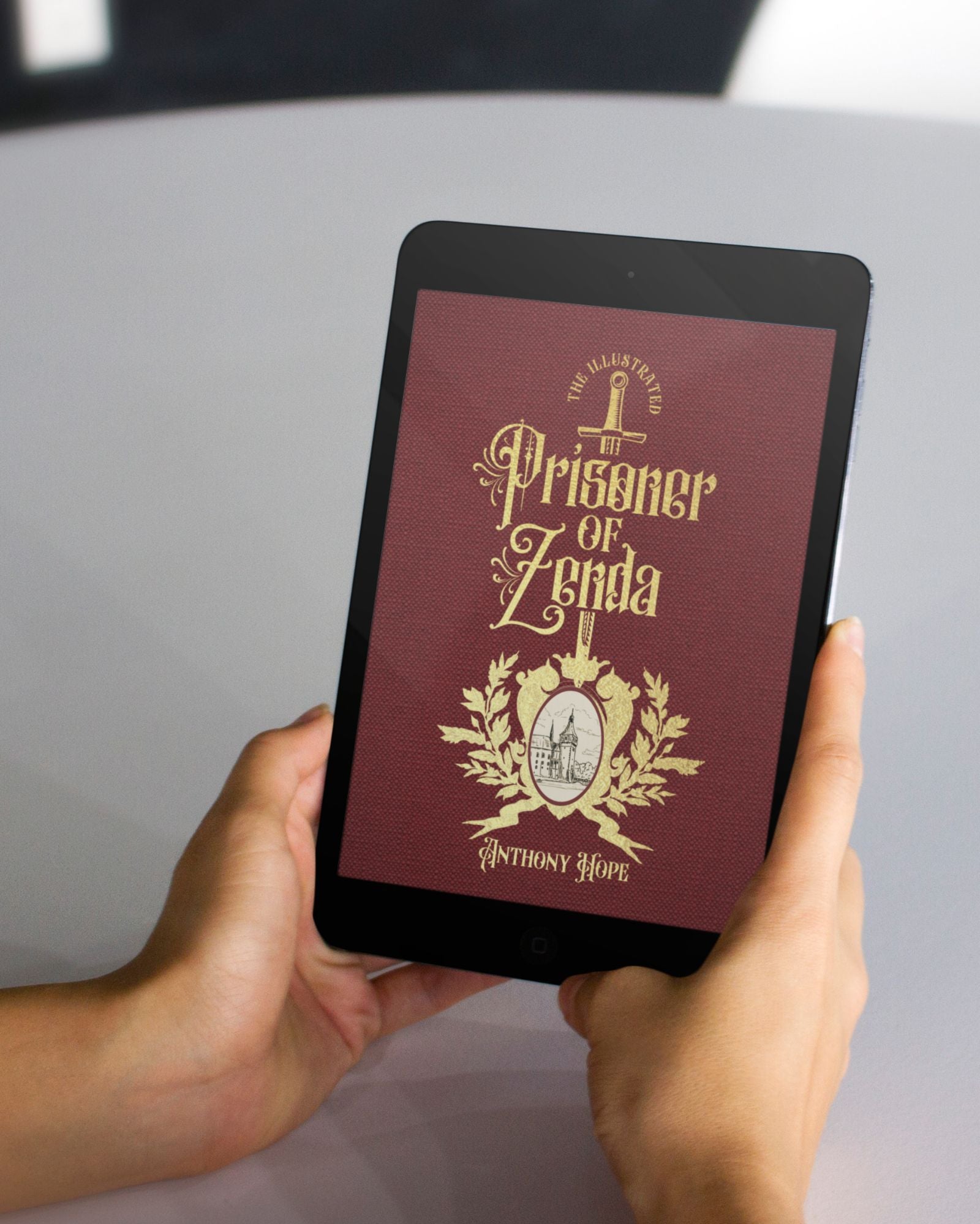 White hands hold a black tablet of the Burgundy and gold ebook "The Prisoner of Zenda" in front of a grey table.