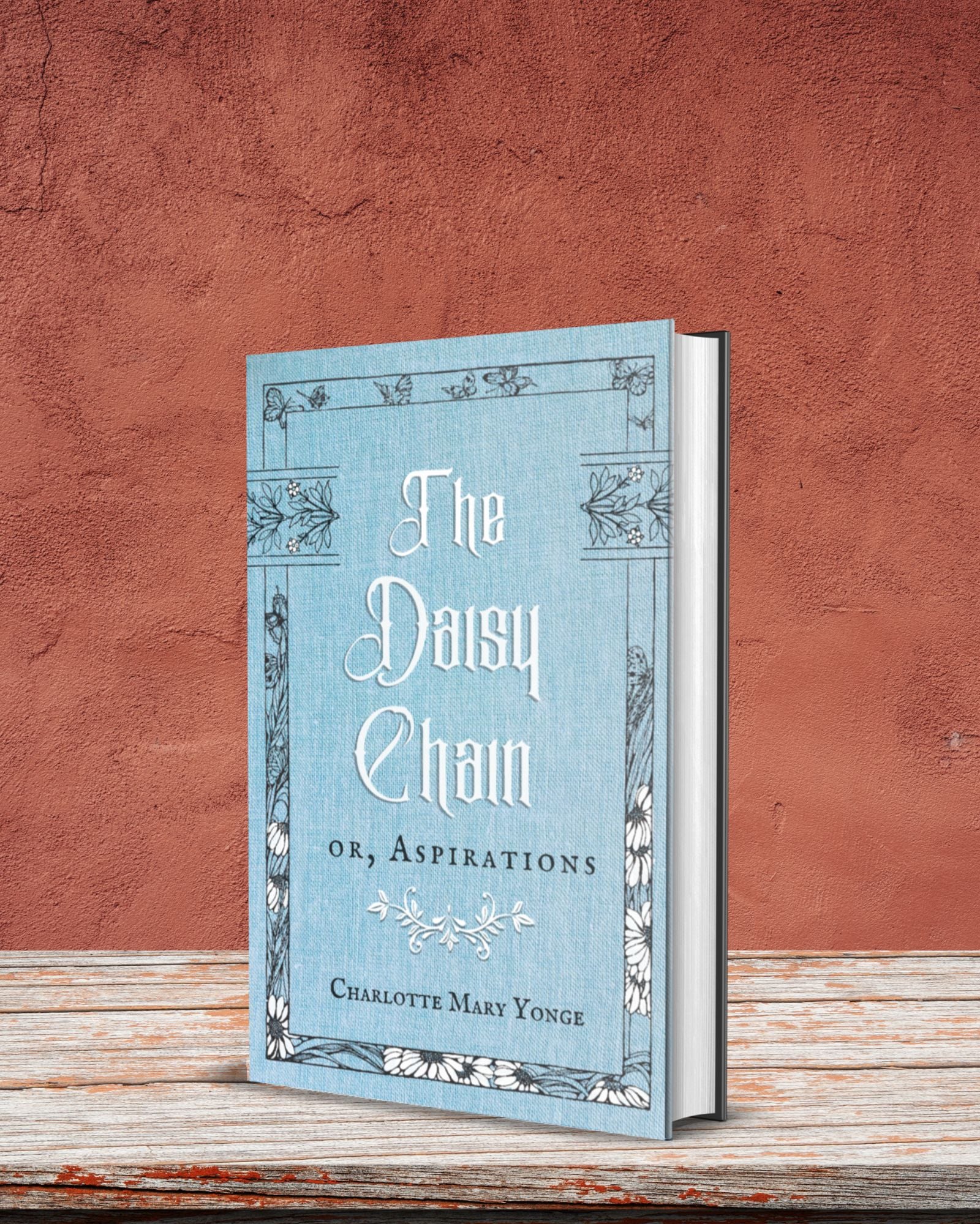Hardback edition of the lovely pale blue and white book "The Daisy Chain or, Aspirations" stands on a rustic table in front of a clay colored wall.