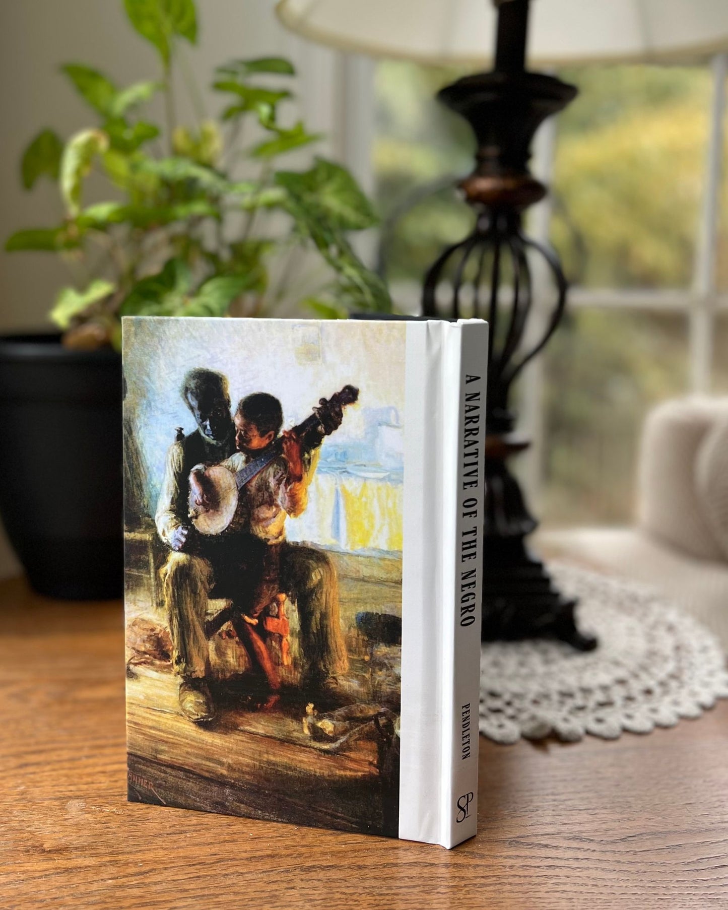The back of the special hardback of A Narrative of the Negro has a famous painting of an older black man teaching a young brown-skinned boy to play the banjo. The book stands in front of a blurred background including a lamp on a doily and plant.