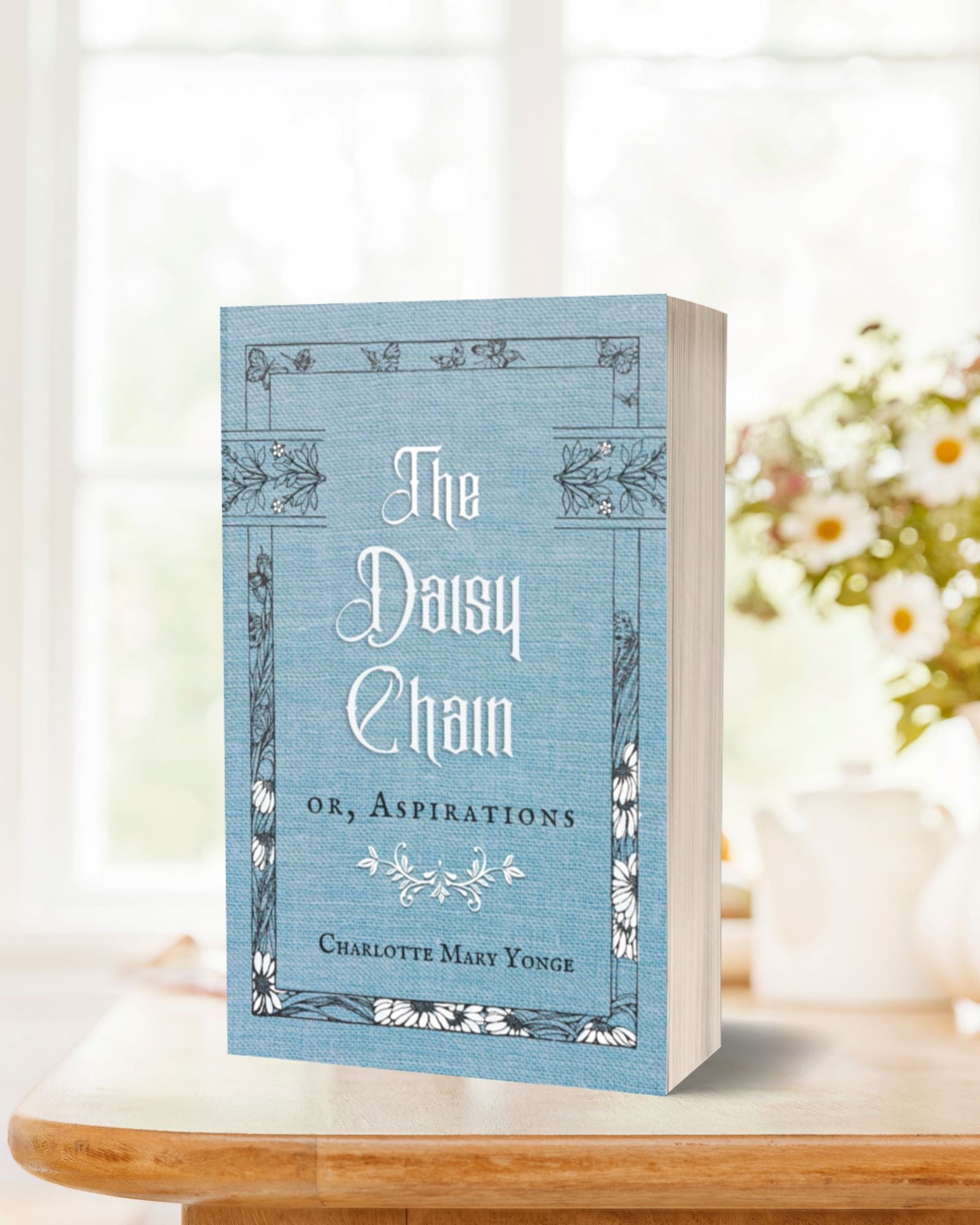 The massive paperback edition of the blue and white book, "The Daisy Chain or, Aspirations" standing on a light table in front of a blurred white background.