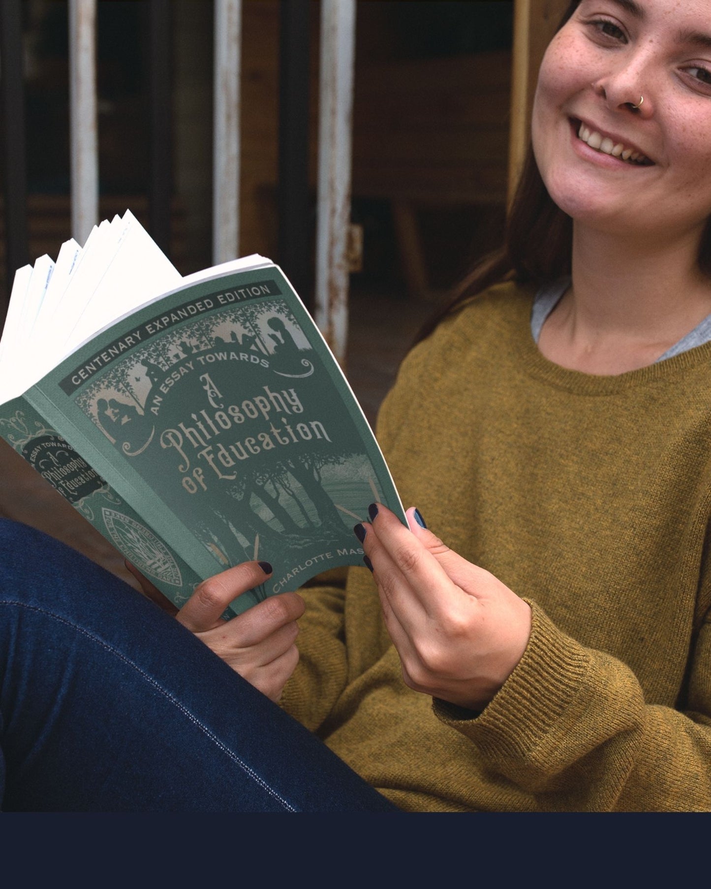 A smiling white girl in jeans and a gold sweater smiles while holding open the paperback edition of the sage colored An Essay Towards A Philosophy of Eduction.